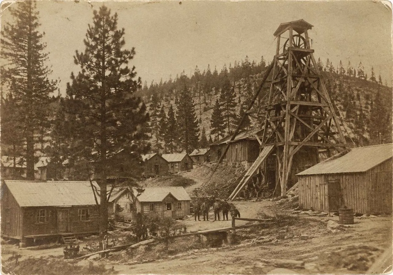 Archival-style photograph of an early 1900s gold mine headframe and bunkhouses among pines